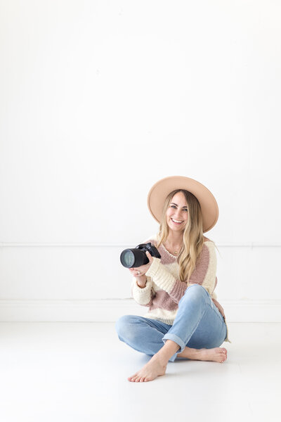 Photographer Amanda Zabrocki smiles in a hat and white blouse in front of white brick wall