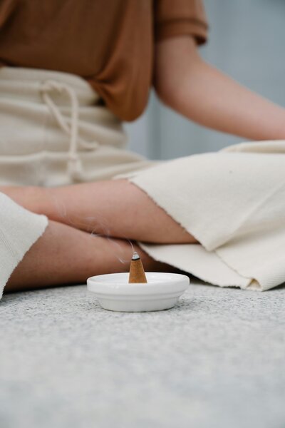 Woman sitting cross-legged in cream pants, with a lit incense cone on the floor in front of her.