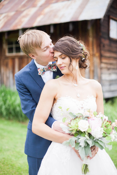 Rustic wedding photography featuring a bride and groom kissing in front of a barn.