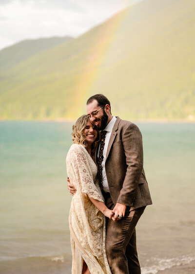 A couple embraces on the shore of a lake, a rainbow beaming down behind them  during their olympic national park wedding