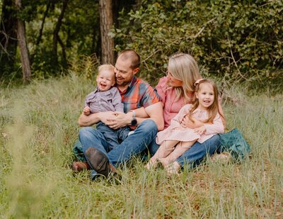 Family sitting in a grassy field sharing laughter and cuddles during a relaxed outdoor portrait session by a Colorado Wedding and Portrait Photographer.
