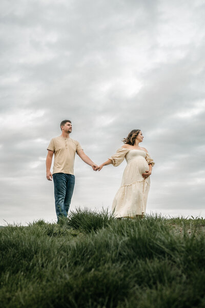 Husband wraps his arms around his expecting wife while they stand in a lush green field. 