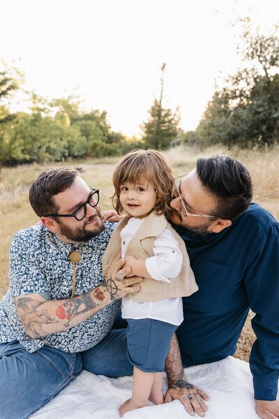 two dads kissing toddler boy in a field at sunset