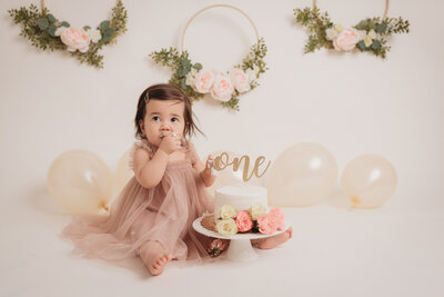 Smash cake portrait session at Atlanta portrait studio with brown haired one year old girl sitting eating her cake with balloon and flower decorations in background