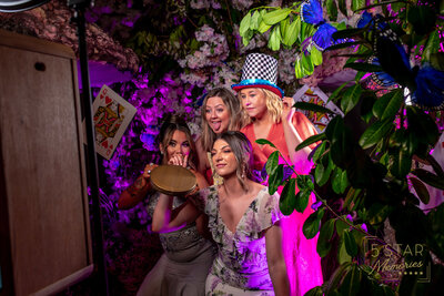 Lady wearing blue dress smiling, a man wearing suit and tie, and the other lady holding a bouquet touches the photo booth screen getting ready to have their photos taken