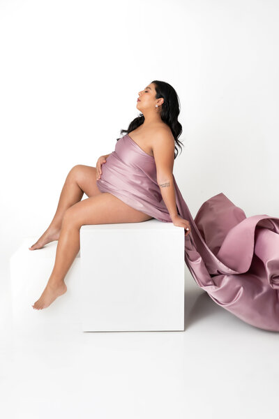 A beautiful mother draped in silky mauve fabric. posing on a white box.
