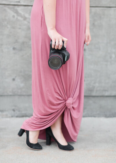person in a pink dress and black heels holding a camera with one hand