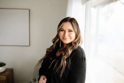 A person with long wavy hair, wearing a black top, stands near a window with sheer curtains. They smile at the camera, capturing the perfect moment for a Minnesota photographer. A blank picture frame hangs on the wall behind them to the left.