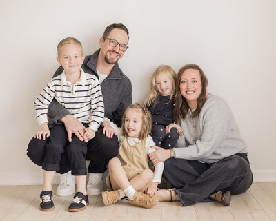 family of five sitting against a wall in a studio smiling at each other and the camera.  Mom, dad, a young son, and two young daughters.  Dad is wearing glasses, with short hair, a goatee, and his son on his lap.  Son is wearing a striped shirt and pants.  Mom has medium length brown hair, wearing a gray sweater, and black pants.  One daughter has short brown hair, wearing a light tan dress and shoes, and white long sleeve shirt.  Youngest daughter is wearing a dark blue and white pants and long sleeve shirt.