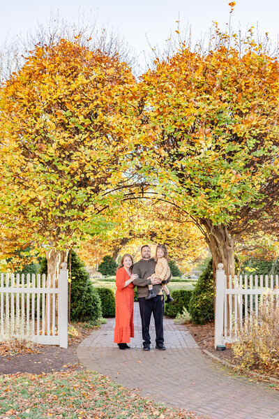 Mom, dad with dad holding daughter in his arms smiling at the camera under a beautiful fall tree arch in Dayton, Ohio at Wegerzyn Gardens.