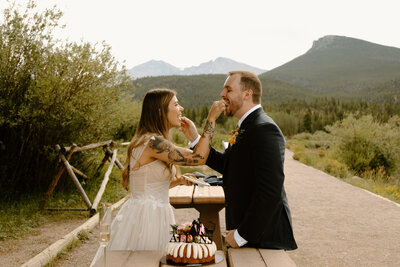 bride and groom feeding each other cake while standing outside with mountains in the background