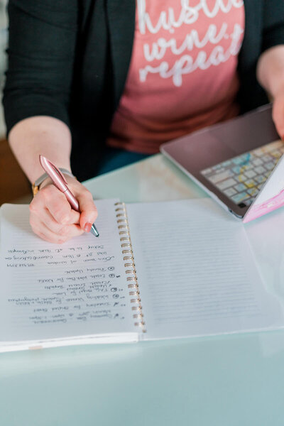 Becca Mai writing in a notebook sitting at a desk on her laptop