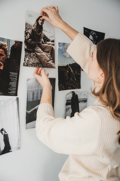 Female photographer is working on a moodboard, by pinning magazine editorial photos to a wall as proces of the pre-production