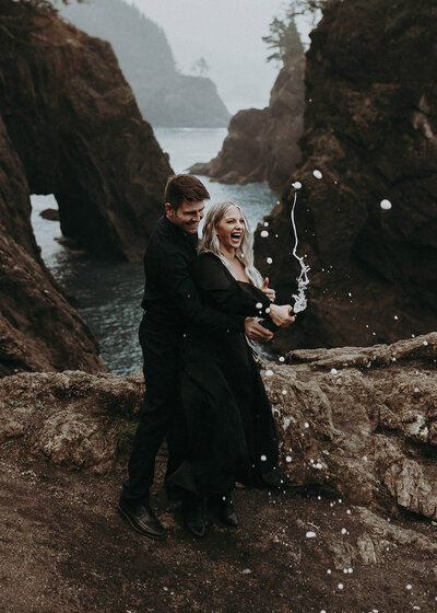 An elopement couple in all black popping champagne in the Samuel H Boardman Scenic Corridor in Southern Oregon.