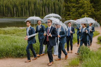 Groomsmen walking with umbrellas in the rain. Overcast photography highlights vibrant, true to color greens and natural Colorado landscape. 