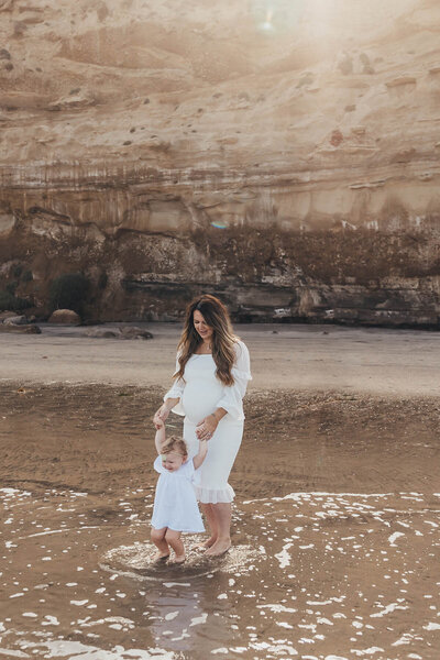 a pregnant mother holds her daughter's hands while they walk in the water at La Jolla Shores beach in San Diego