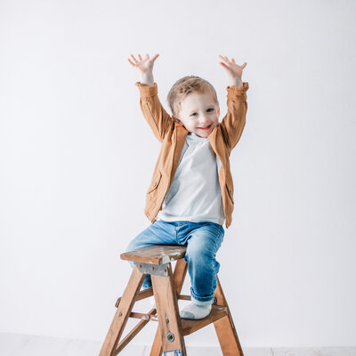 little boy on a small ladder smiles a the camera with his hands up in the air showing how big he is