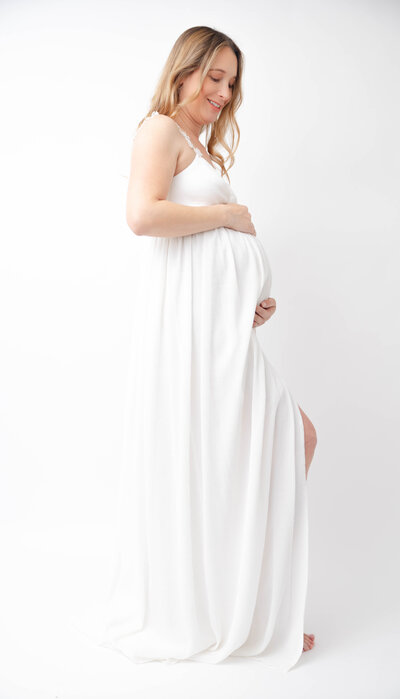 Pregnant mom wearing a white long dress with hands placed above and bellow belly.
