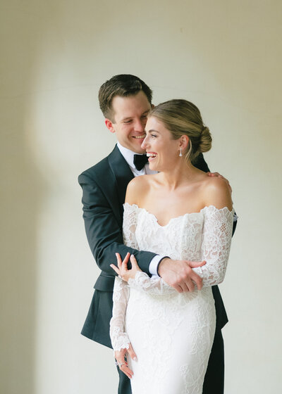 Bride and groom smiling at each other outside with their wedding venue in the background