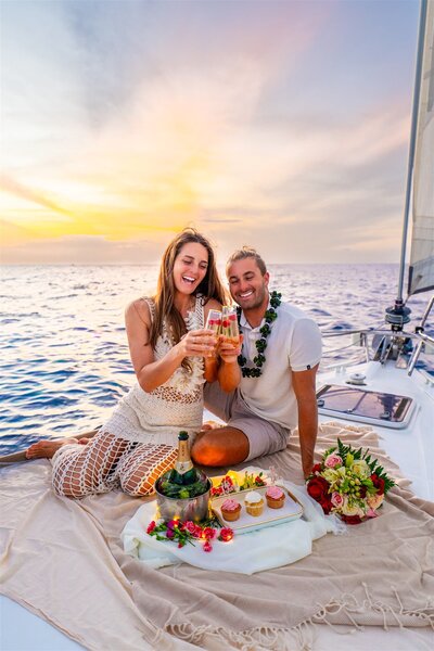 Engaged couple sitting on a sailboat in Hawaii toasting with drinks at sunset, with cupcakes, champagne, and a bouquet arranged beside them.