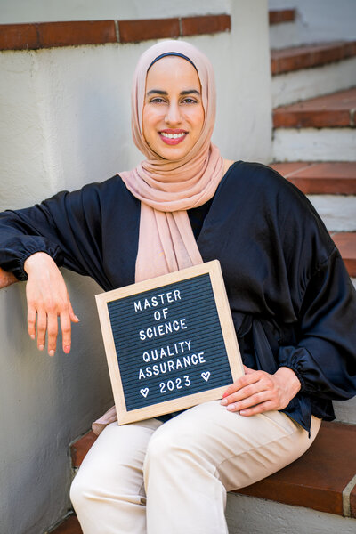 A smiling young woman in a hijab sits on a white and brick staircase with her arm propped up on the banister, and her lap holding a sign that reads "Master of Science, Quality Assurance, 2023" Photo by SAVI Photography - Los Angeles CA Photographer