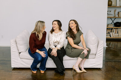 Three women sit on a white couch in a relaxed setting. They are smiling and laughing, dressed in casual and semi-formal attire. The background features a white wall and a shelf with books and decorative items.