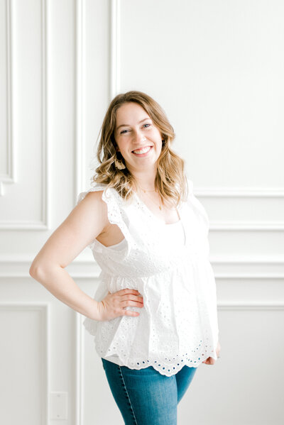 A headshot of Cleveland Family & Newborn Photographer, Brittany Serowski Photography owner, Brittany Serowski. Standing in front of an all white wall at the Lake Arts Studio in Avon Lake, OH, she is wearing dark blue jeans and a white tank top as she smiles walking towards the camera.