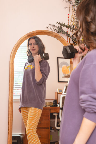 Photo of Cassie Paton lifting a dumbbell while looking at her reflection in a mirror, wearing a purple sweatshirt and yellow checkered leggings in a cozy, plant-filled home office. - Mettle & Tonic