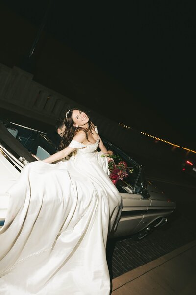 Bride in off-the-shoulder gown smiling while sitting on vintage car at night.
