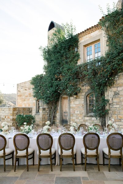 An outdoor dinner table stretches across a courtyard, framed by a stone villa and lush greenery.