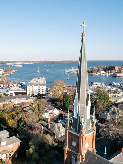Aerial view of a coastal town with a tall church steeple in the foreground. Sailboats are on the water, and buildings and houses surround the area. The sky is clear and blue.