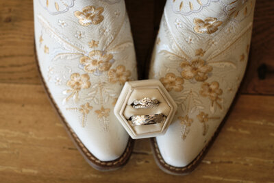 Top down photo of a pair ofWhite cowboy boots with rose gold embroidery. On the toes of the boots is a ring box with the couples wedding bands inside.