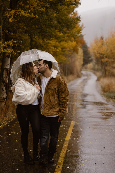 A couple kisses while standing under a clear umbrella on a wet mountain road, surrounded by golden autumn trees.