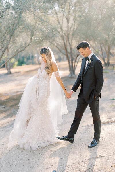 A bride and groom walk hand in hand, her dress is lace a toile, in a mermaid style, she is holding onto her long trailing veil.