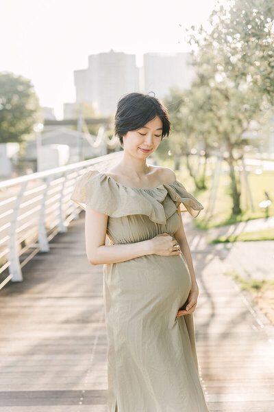 A pregnant woman holding her belly with both of her arms and looking down in her green khaki dress. Maternity photo session in Tokyo Japan by Yukika Ishikawa Photography.