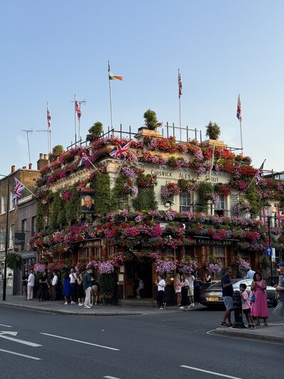 pub in London near earls court with flower baskets and greenery encasing the building, people mingling on the sidewalk with pints and flags on top waving in the breeze