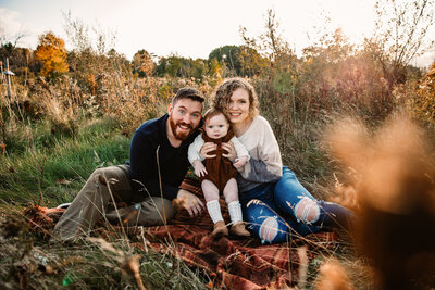 mom dad and baby smile at camera in a field on a blanket at Nashotah park