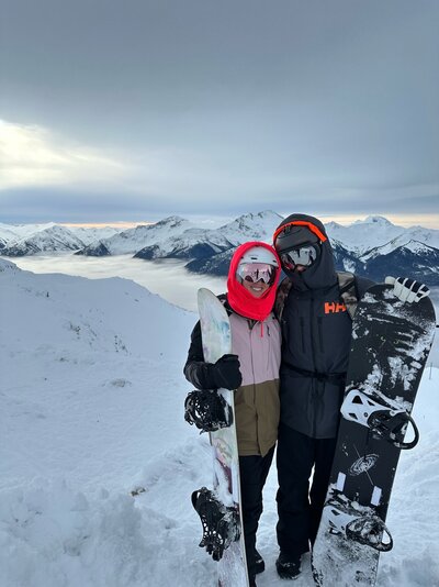 Emily and her husband stand on the top of a mountain holding snowboards