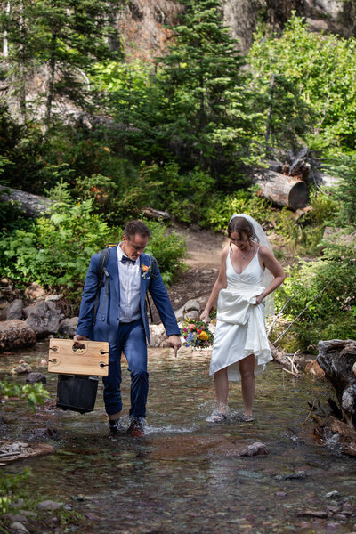 Bride and groom wade barefoot through a clear mountain stream, carrying a picnic setup in the forest.