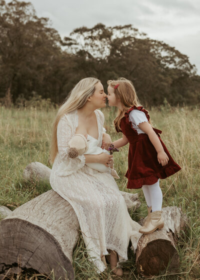 Big sister going in to kiss her baby sister that is being held by dad while mum watches on with her arms around her daughter.