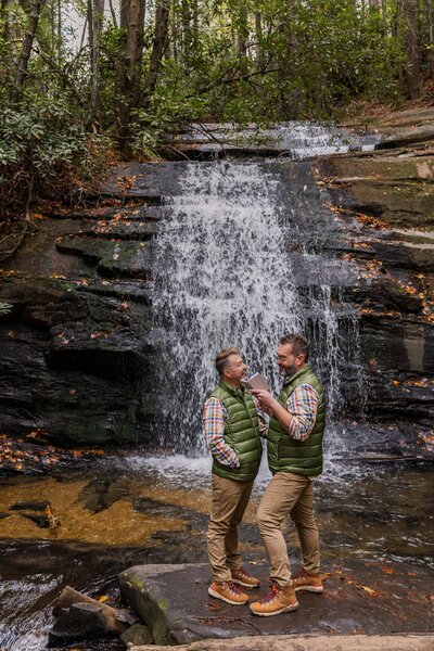Two grooms stand on a flat rock in front of a large cascading waterfall. The waterfall is falling into a natural pool behind them. They are standing facing each other while reading vows and looking lovingly at one another. 