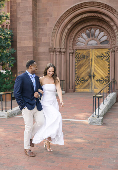 A couple walks arm-in-arm in front of an ornate, wooden door set in a brick building. The man wears a navy blazer and tan pants, while the woman is in a strapless white dress, smiling as she looks at him. They are surrounded by greenery.