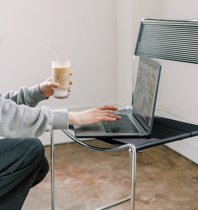 Side view of a woman typing on a MacBook Pro balanced on a modern chair while holding an iced coffee.