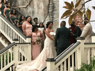 A bride and groom stand facing each other during their wedding ceremony on an outdoor staircase, surrounded by bridesmaids in pink dresses and groomsmen, with an officiant and lush greenery nearby.