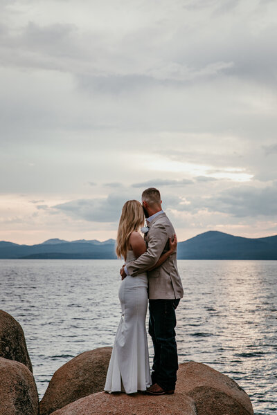 Couple embracing on the rocky shoreline at Sand Harbor in Lake Tahoe, with soft evening light reflecting on the water and mountains in the distance.