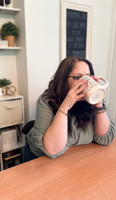 Ali sipping from "Sun Please" mug at her desk with shelves and decor in the background