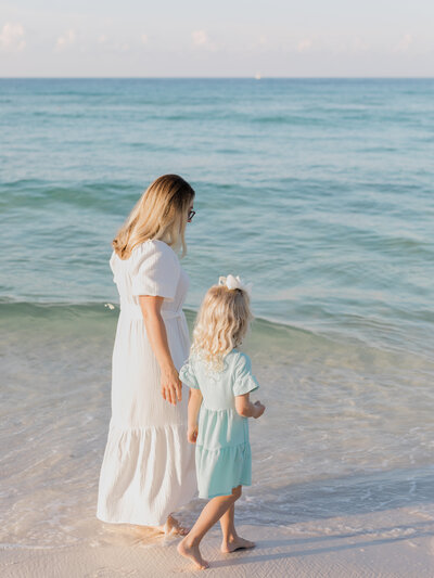 A mother in a white dress walking along the 30A shore in the turquoise ocean water with her blond, curly haired daughter in a light blue dress.