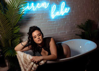 Boudoir Photographer, a woman wears sexy attire and kneels in a tub with neon lihhts behind her that read "siren babe"