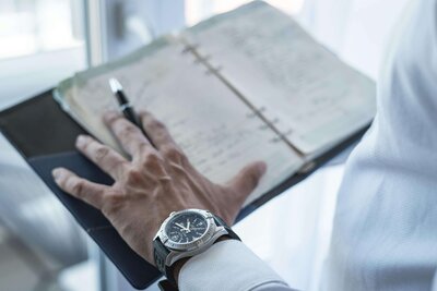 A man's hand resting on a desk calendar, holding a pen, with a blurred office background.