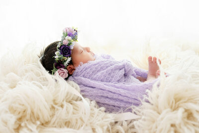 Baby wrapped in a lilac fabric, laying on a flokati and a headband with different shades of purple flowers.
